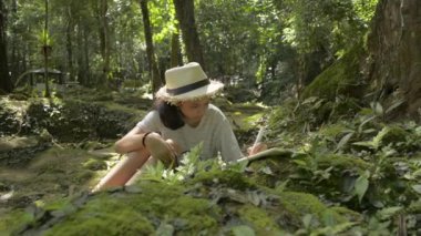 Hasır şapka takan Asyalı genç bir kız oturmuş ve tropikal ormanların koruma için doğası hakkında not alıyor. Sınıf dışından bilgi. Döndürme çekimi.