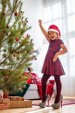 İyileşir yüksek üzerinde duran küçük kız ayakkabı ve parlak kırmızı elbise Noel ağacı ve gülümseyerek elini kadar yükselen
