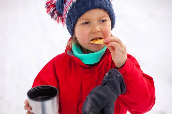 Крупный план портрета маленькой девочки в красной зимней куртке, пьющей чай из термоса с печеньем — стоковое фото