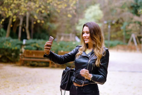 Charming young female in trendy outfit smiling and taking selfie while standing on blurred background of park trees