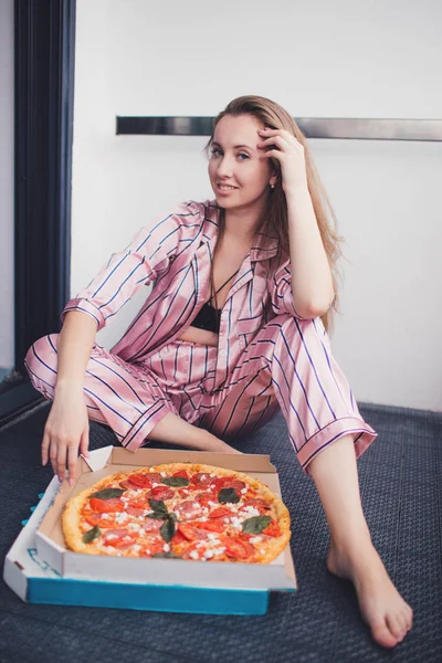Ung kvinna äter en bit pizza sittandes på golvet i pyjamas. — Stockfoto