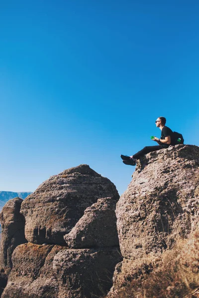 Reppupäinen kaveri istuu vuorilla kivellä, juo teetä ja nauttii näköalasta luontoon. . — kuvapankkivalokuva