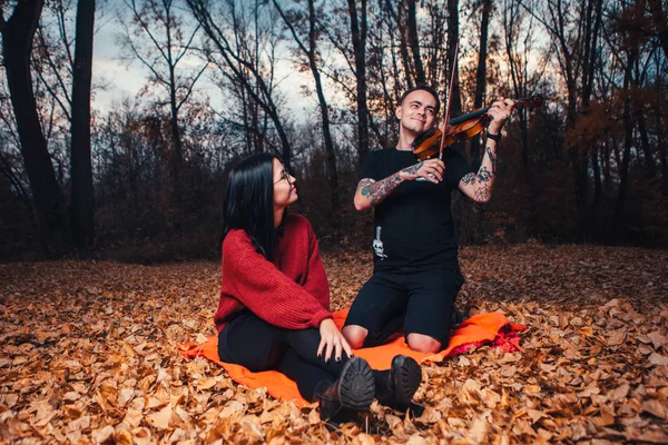 Young man play on violin and young woman are sitting on a plaid in an autumn forest — Stock Photo, Image