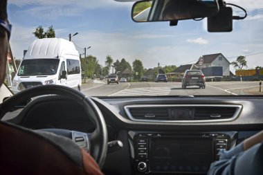 Otomobilin açık penceresinden iki yönlü trafik ve geçen arabaların üstünde o ile görünür geniş yol var. Yolu ile küçük bir köy geçer.