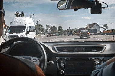 Otomobilin açık penceresinden iki yönlü trafik ve geçen arabaların üstünde o ile görünür geniş yol var. Yolu ile küçük bir köy geçer.