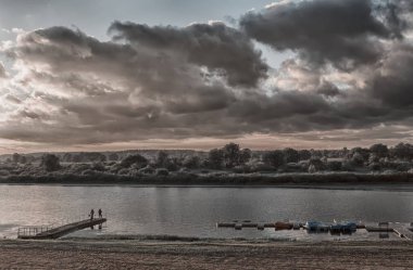 Ahşap iskelesi olan küçük bir nehir batan güneşle aydınlanır. Gökyüzü karanlık ve fırtınalı..