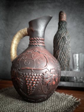 El yapımı örgülü kırmızı kilden yapılmış seramik şarap sürahisi. Yanında bir şarap şişesi ve bir bardak var..