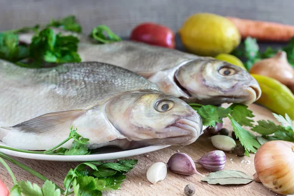 皿の上に2つの大きな鯛があります 近くにはこの川魚を調理するための野菜やスパイスがあります フロントビュー クローズアップ — ストック写真