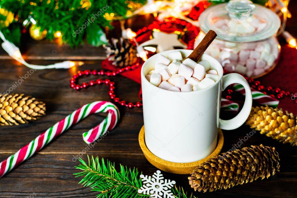 White mug of cocoa with marshmallows, lollipops, fir and pine cones, Christmas tree branch with garland and snowflake on brown wooden table, Christmas mood background