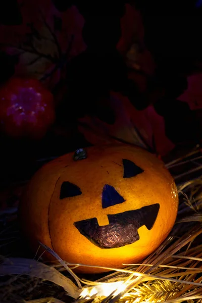 Single funny or angry pumpkin Jack o\'Lantern head in the hay, against the background of autumn yellow leaves, the light shines in his face. On a dark wooden board, vertically, copy space