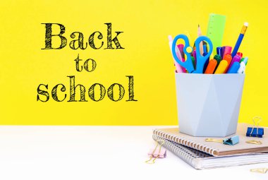 Eğitim konsepti. Not defterleri, kırtasiye ve düzenleyici çok renkli kalemler, kalemler, kalemler ve makaslar beyaz bir masa üzerinde sarı bir zemin üzerinde yazıyla okula geri dönüyor..