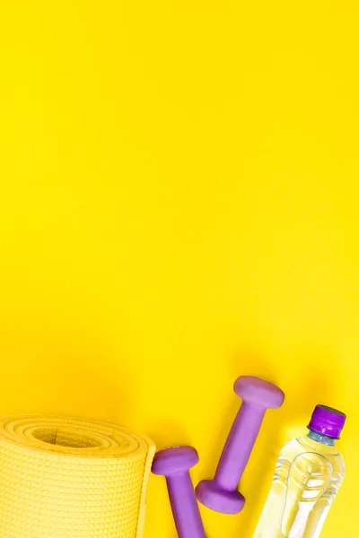 Home fitness concept yellow yoga mat, two purple dumbbells and a bottle of water. Equipment for training and exercises for weight loss. Flat lay a bright vertically background with copy space.