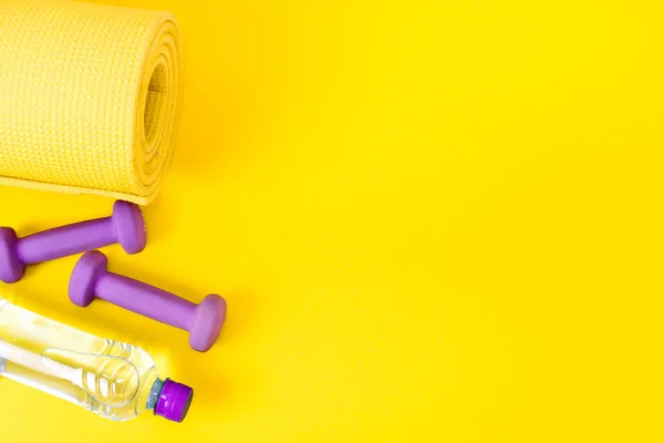 Home fitness concept yellow yoga mat, two purple dumbbells and a bottle of water. Equipment for training and exercises for weight loss. Flat lay a bright background with copy space.