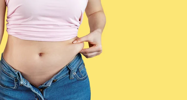 Woman in unbuttoned jeans holds with hand and squeezes excess belly fat. The concept of overweight, weight loss, diet, obesity, junk food on yellow background, banner, copy space.