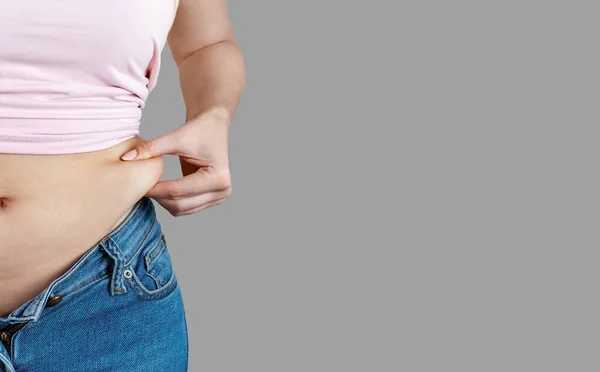 Woman in unbuttoned jeans holds with hand and squeezes excess belly fat. The concept of overweight, weight loss, diet, unhealthy lifestyle, obesity, junk food on gray background, banner.