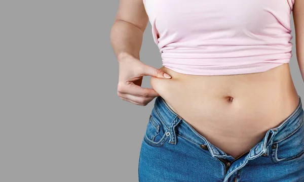 Düğmesiz kot giyen kadın elini tutar ve göbek yağlarını sıkar. Aşırı kilo verme, zayıflama, diyet, sağlıksız yaşam tarzı, obezite, gri arka planda abur cubur, afiş.