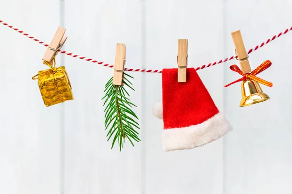 Santa Claus red hat, jingle bell, Christmas tree branch and gift box hangs on a rope and is attached with a wooden clothespins. Traditional symbols of the New Year on a light background.