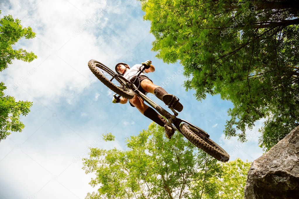 Young cyclist flying with his bycicle from a rock in the forest. Extreme low angle view