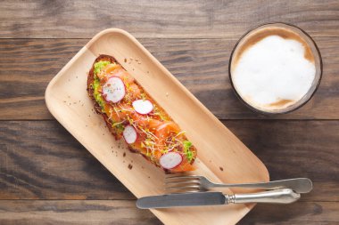 Rustik ekmek tost püresi avokado, somon füme, turp, brokoli ile lahanası ve tohum rustik ahşap bir masa üzerinde sütlü bir kahve ile ahşap bir tabak içinde görev yaptı. Üstten görünüm, düz yatıyordu