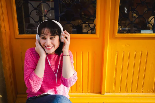 美丽和开朗的年轻女子听音乐与耳机和智能手机出一个商店的黄色门 — 图库照片
