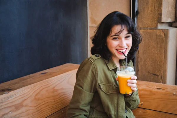 屋外ストローで果物のスムージーを飲む緑色のジャケットを持つ幸せな女 — ストック写真