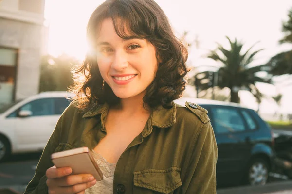 Young beautiful woman with green jacket using her mobile phone in street at sunset looking to camera and smiling