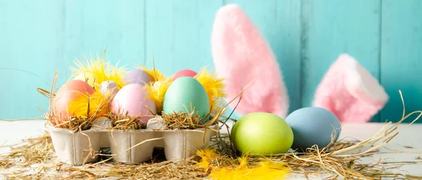 Pastel colored easter eggs in a egg box with hay and feathers as a nest and rabbit ears behind against a wooden turquoise background. Banner size