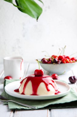 İtalyan panna cotta tatlısı, kiraz soslu beyaz ahşap masa ve üstünde canavar yaprağı.