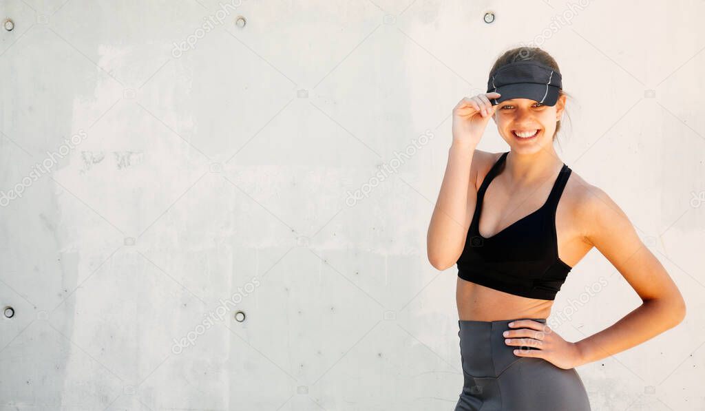 Portrait of a beautiful and happy sporty young woman with stylish sportswear and a visor cap posing against a white wall in the street. Copy space