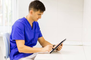 Doctor wearing blue coat working on line with a tablet in a dentist clinic. clipart