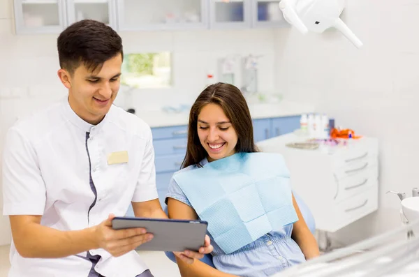 Молодий стоматолог в стоматологічному кабінеті розмовляє з пацієнткою і готується до лікування . Стокова Картинка