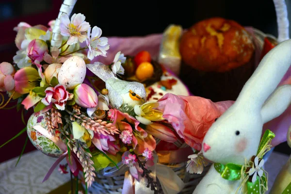 Paskalya kompozisyon. Çiçeklerle süslenmiş, boncuk, yumuşak bir beyaz tavşan yanındaki bir Paskalya sepeti bir kuş ve bir yumurta yaptı. Arka arka plan - Paskalya kek bulanık.