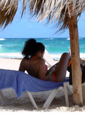 Punta Cana, Dominik Cumhuriyeti, Aralık 2018. Saman şemsiyesi altında sahilde güzel bir siyah kız kıvırcık saçlı ve elleri bir telefon sunbathes.