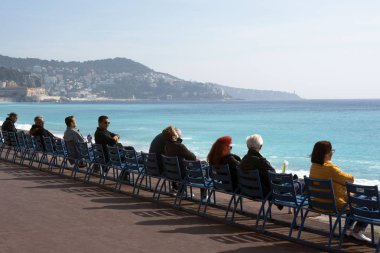 Nice, France, March 2019. People sit on the famous blue chairs on the Promenade des Anglais, watch the azure sea and enjoy the warm sunshine. Rest and relaxation on the Cote d'Azur.