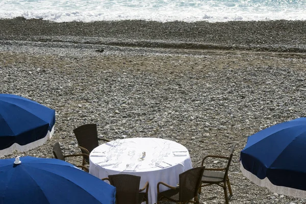 Blue umbrellas, reserved tables with white tablecloths on the pebble beach of the Promenade des Anglais in Nice, France, await guests. Cozy restaurant on the Cote d\'Azur.