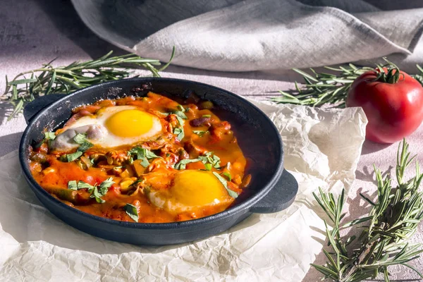 Shakshuka es un sabroso plato de huevos, frito en una salsa de tomates, ají, cebolla y condimentos en una sartén de hierro fundido negro. Tomate, sartén, servilleta y romero en los rayos del sol de la mañana . — Foto de Stock