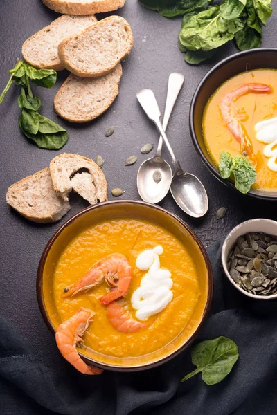 2 dishes of orange pumpkin soup on a black table. Three red shrimps decorate the soup. Spinach, pumpkin seeds and croutons lie on the table. Delicious dinner. Beautiful presentation of dishes. — Stock Image 2 dishes of orange pumpkin soup on a black table. Three red shrimps decorate the soup. Spinach, pumpkin seeds and croutons lie on the table. Delicious dinner. Beautiful presentation of dishes. Royalty Free Stock Photos