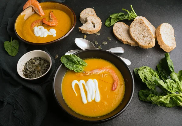 2 dishes of orange pumpkin soup on a black table. Three red shrimps decorate the soup. Spinach, pumpkin seeds and croutons lie on the table. Delicious dinner. Beautiful presentation of dishes. — Stock Image 2 dishes of orange pumpkin soup on a black table. Three red shrimps decorate the soup. Spinach, pumpkin seeds and croutons lie on the table. Delicious dinner. Beautiful presentation of dishes. Stock Picture