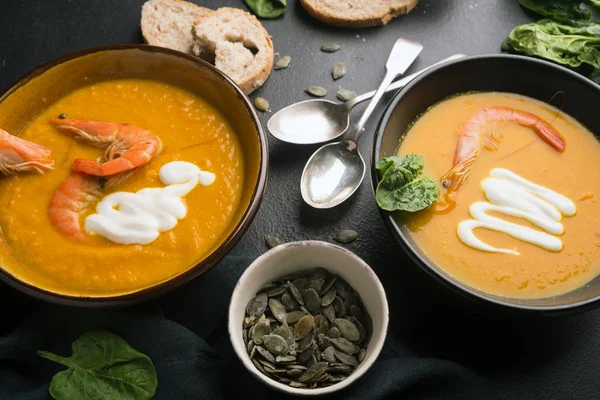 2 dishes of orange pumpkin soup on a black table. Three red shrimps decorate the soup. Spinach, pumpkin seeds and croutons lie on the table. Delicious dinner. Beautiful presentation of dishes. — Stock Image 2 dishes of orange pumpkin soup on a black table. Three red shrimps decorate the soup. Spinach, pumpkin seeds and croutons lie on the table. Delicious dinner. Beautiful presentation of dishes. Stock Image
