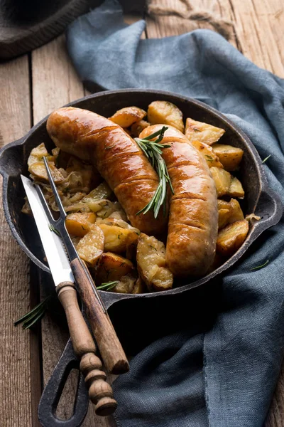 Delicious hot hearty food: fried potatoes with sausages on the grill in a cast iron pan. Knife and fork with wooden handles. High calorie lunch. Traditional German cuisine.