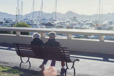 Yaşlı bir çift güneşli bir günde beyaz pahalı yatlar ve dağlar arasında bir bankta oturuyor. Eski yaşta seyahat. Akdeniz 'de yat Parkı, Cannes, Fransa.