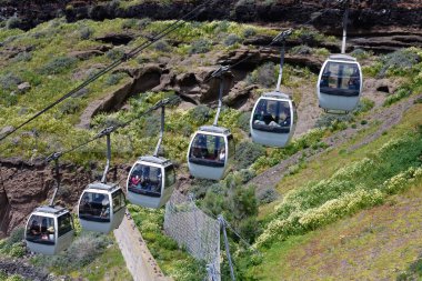 Santorini, Yunanistan, Nisan 2019. Teleferiğin kabinlerinde turistler Santorini eski limanından yükselir. Kır çiçekleri ile kaplı dağların güzel görünümü.