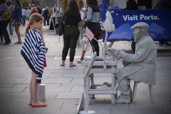 Oporto, Portugal, junio 2019. La niña sostiene su dedo en la boca y mira pensativamente la pantomima de la calle. Estatua viva de un zapatero en el paseo marítimo de Oporto . — Foto de Stock