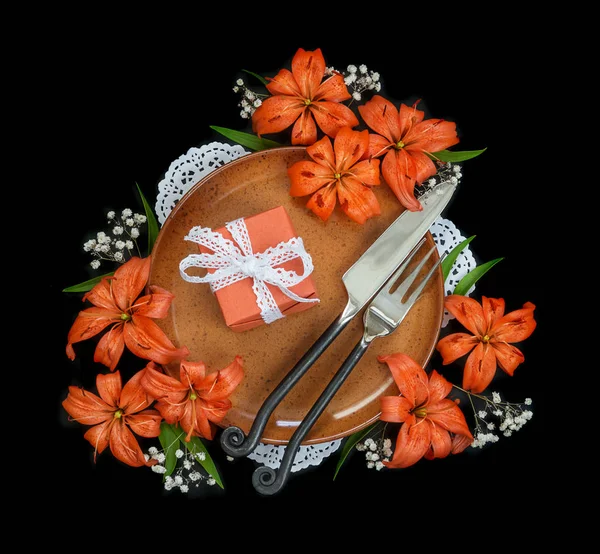 Terracotta plate decorated with orange lilies flowers, gift box tied with white lace ribbon and exclusive forged cutlery isolated on a black background; top view, flat lay