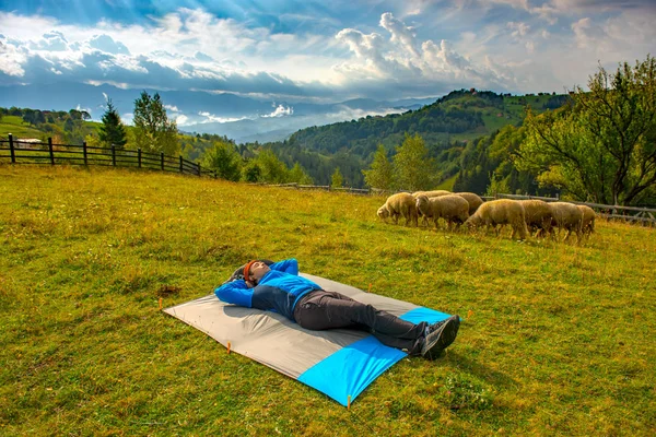 Young mountain hiker resting on a waterproof nylon blanket in a beautiful mountain landscape, while a dozen of sheep are grazing peacefully nearby.