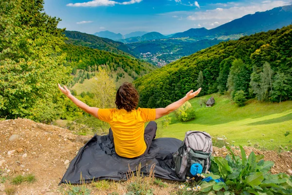 Young mountain hiker sitting on a waterproof nylon blanket in a beautiful mountain landscape and enjoying the view, with arms raised high in the air, celebrating nature and the sunny summer day.