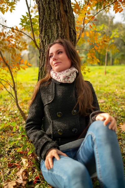 Beautiful romantic girl in a park autumn scenery, sitting down and leaning against a tree, enjoying the warm sunny day. Gorgeous young woman outdoors. Body shot in natural light, vibrant colors