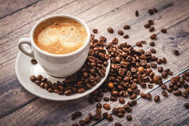 Beyaz bardakta kahve, çay tabağı ve ahşap masa arkasında kahve taneleri..