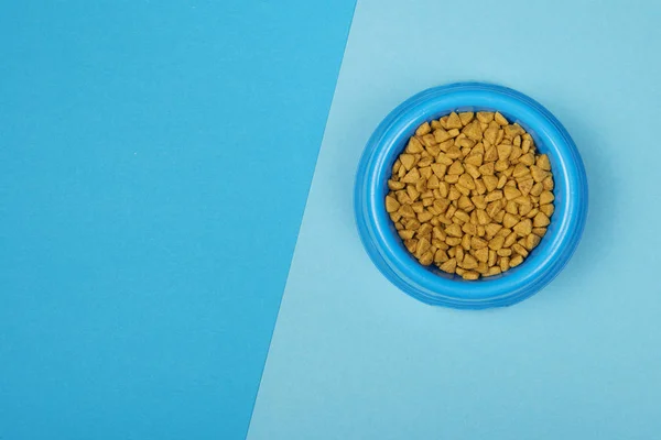 Round Blue food bowl with cat kibble seen from a high angle view on a background of two colors of blue with copy space