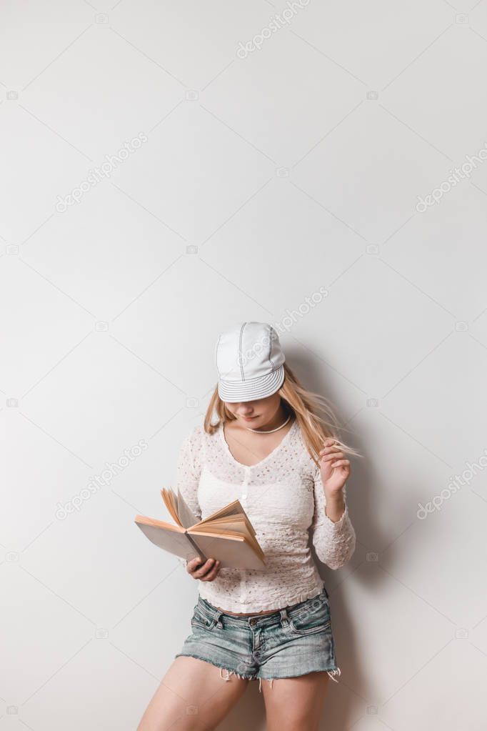 Casual young woman reading a book, leaning against white wall.